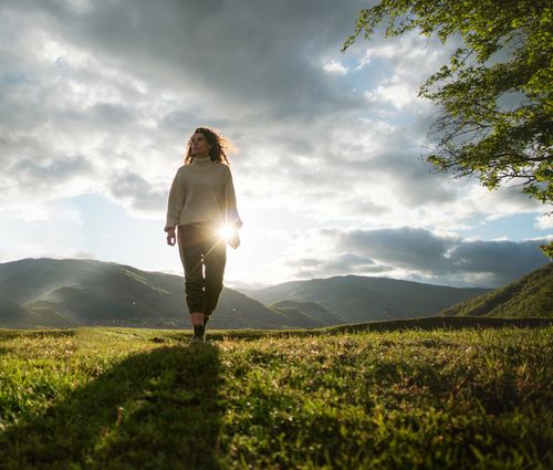 Free A serene woman walks in a sunlit grassland with mountains and clouds in the background. Stock Photo