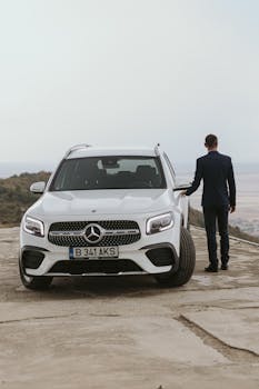 Man in a black suit with a white Mercedes SUV on a scenic mountain road in Romania.