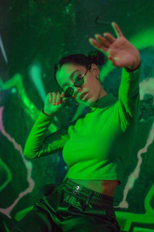 Young Woman in a Casual Outfit and Sunglasses Sitting on the Background of a Wall with Graffiti in Green Lighting 