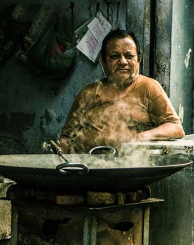 An elderly man cooks in a rustic street market setting with a smoky stove.