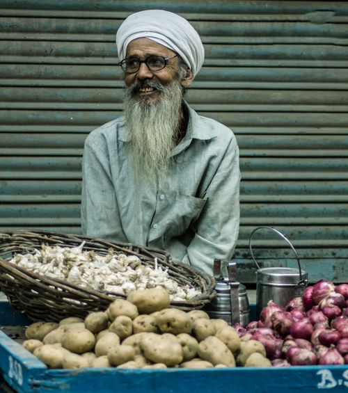 Free Elderly vendor with turban and beard smiling at a produce stall featuring potatoes, onions, and garlic. Stock Photo