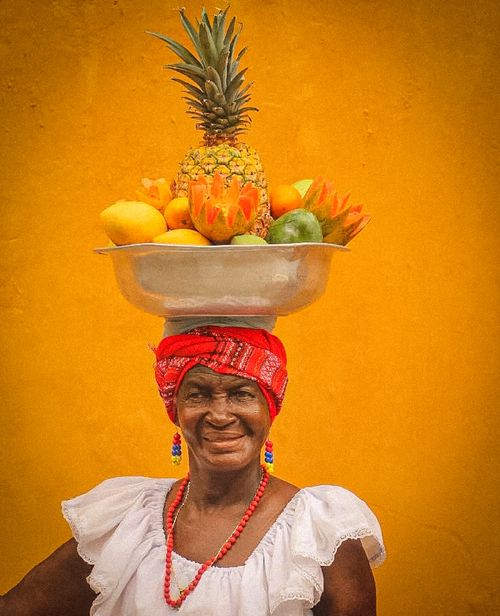 Free Hispanic woman in vibrant attire balancing a bowl of tropical fruits against an orange wall in Cartagena. Stock Photo