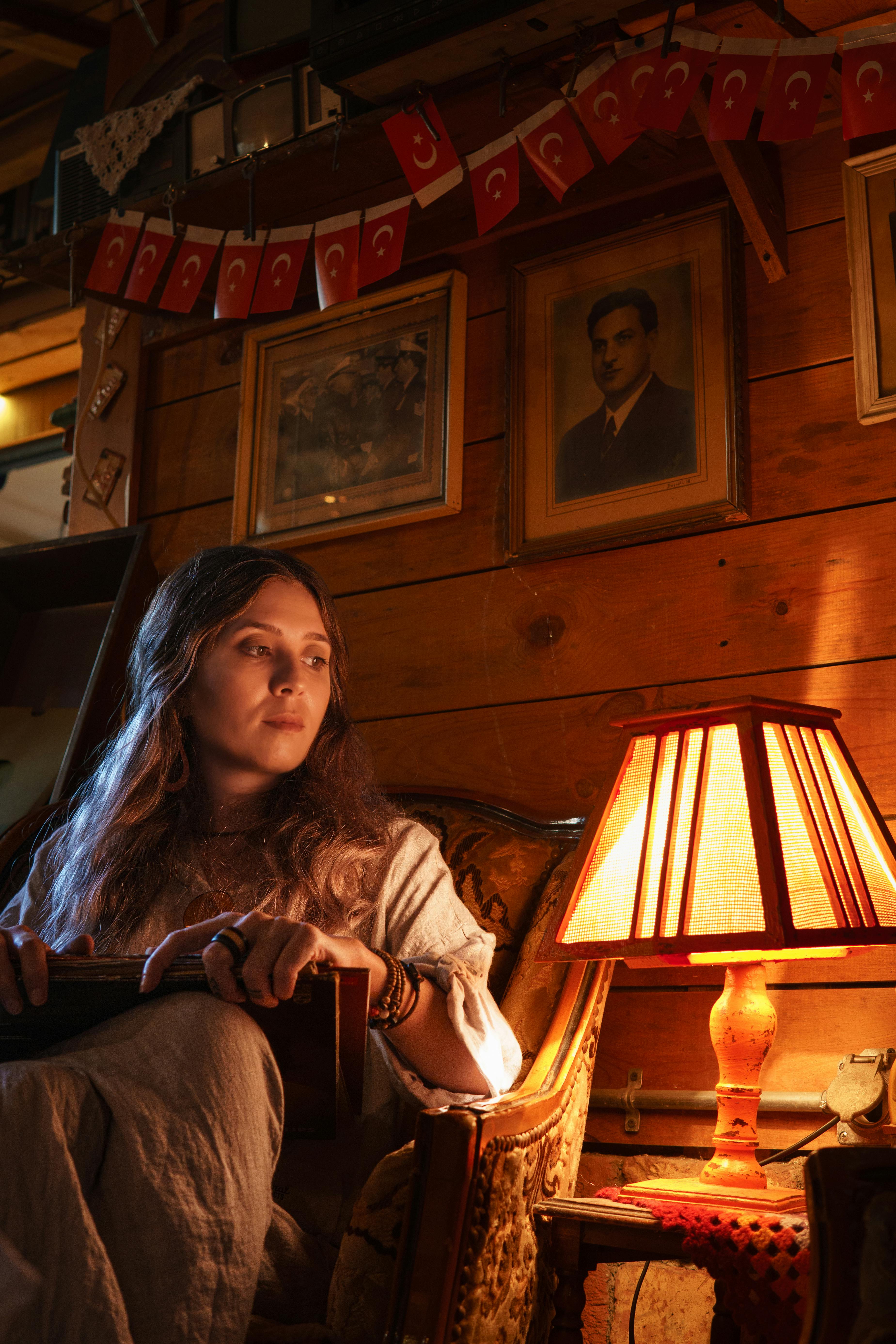Woman sitting in a warmly lit room with a lamp, surrounded by vintage decor and portraits.
