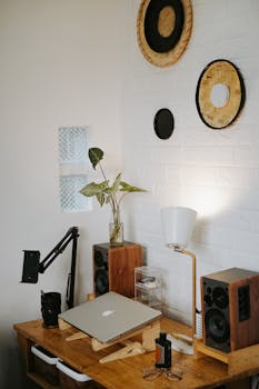 Modern home office desk with laptop, speakers, lamp, and wall decor in a cozy interior.