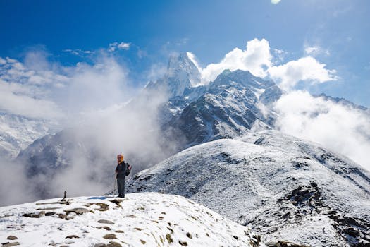 A lone trekker stands atop a snowy mountain ridge in Nepal, with stunning peaks and clouds as the backdrop.