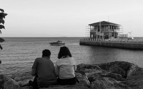 Free A couple sits on rocks by the sea, overlooking a boat and a concrete house. Stock Photo