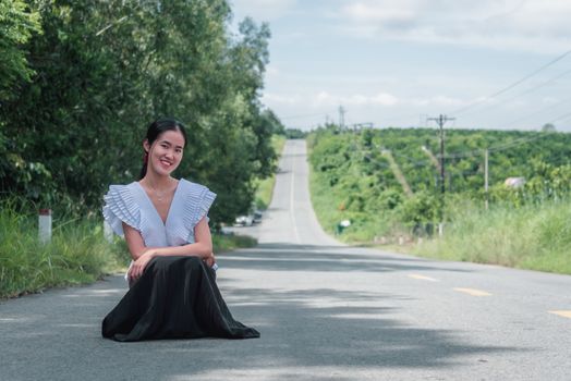 A happy woman in a white top and black skirt sits on a sunny road surrounded by greenery.