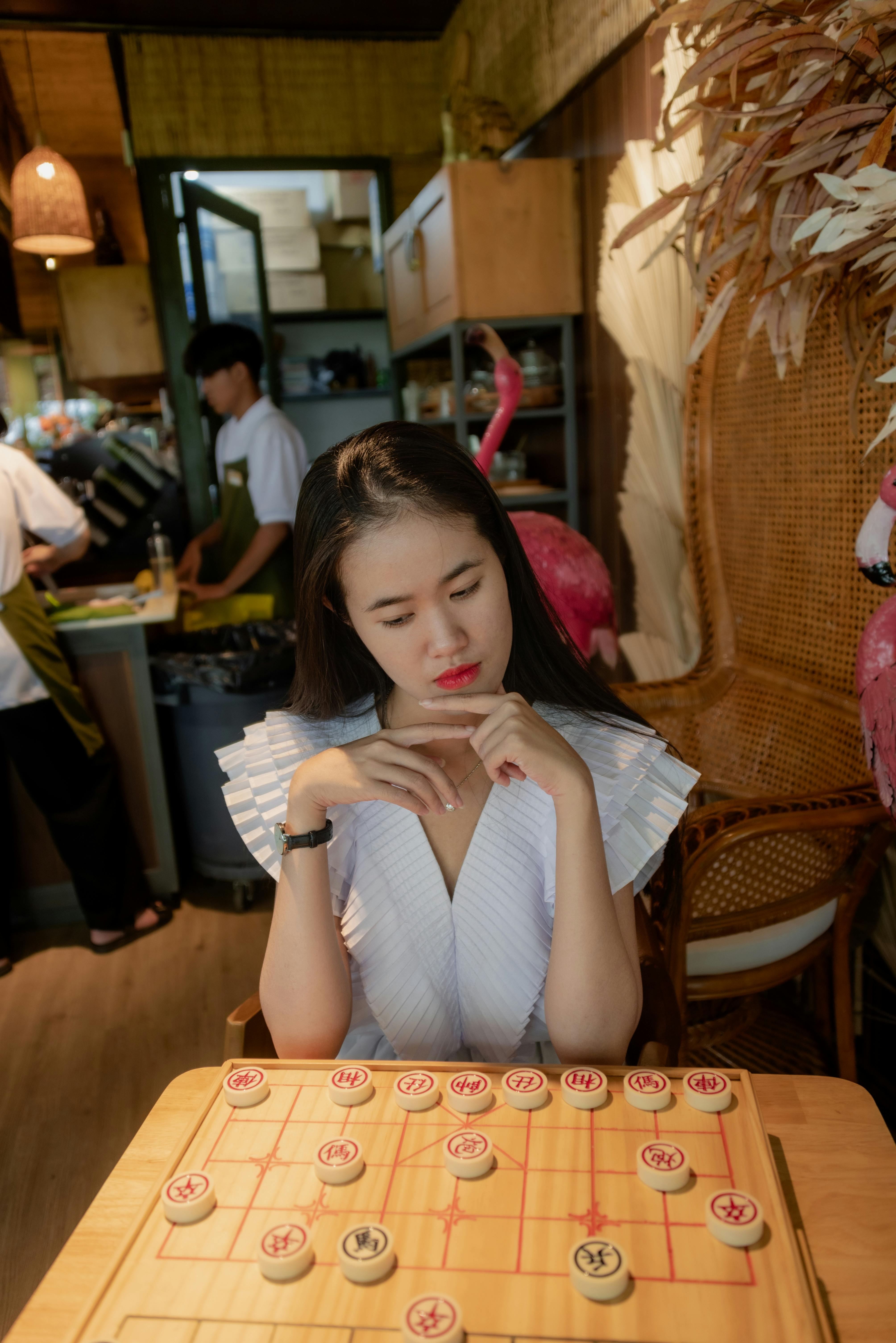 Contemplative Asian woman plays Xiangqi in a cozy cafe. Indoors, thoughtful ambiance.