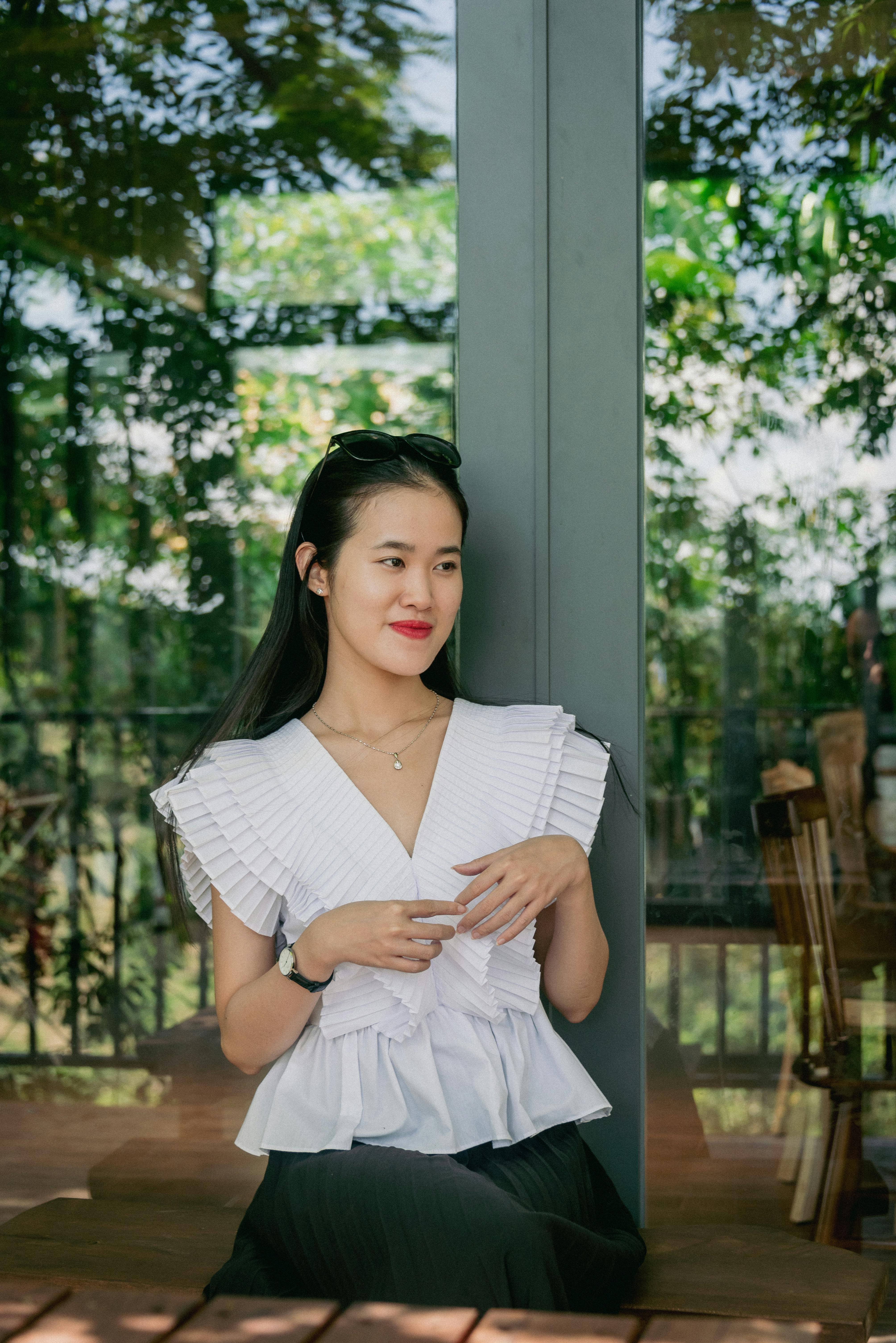 Elegant portrait of a young Asian woman in a white blouse standing by a glass window with greenery i