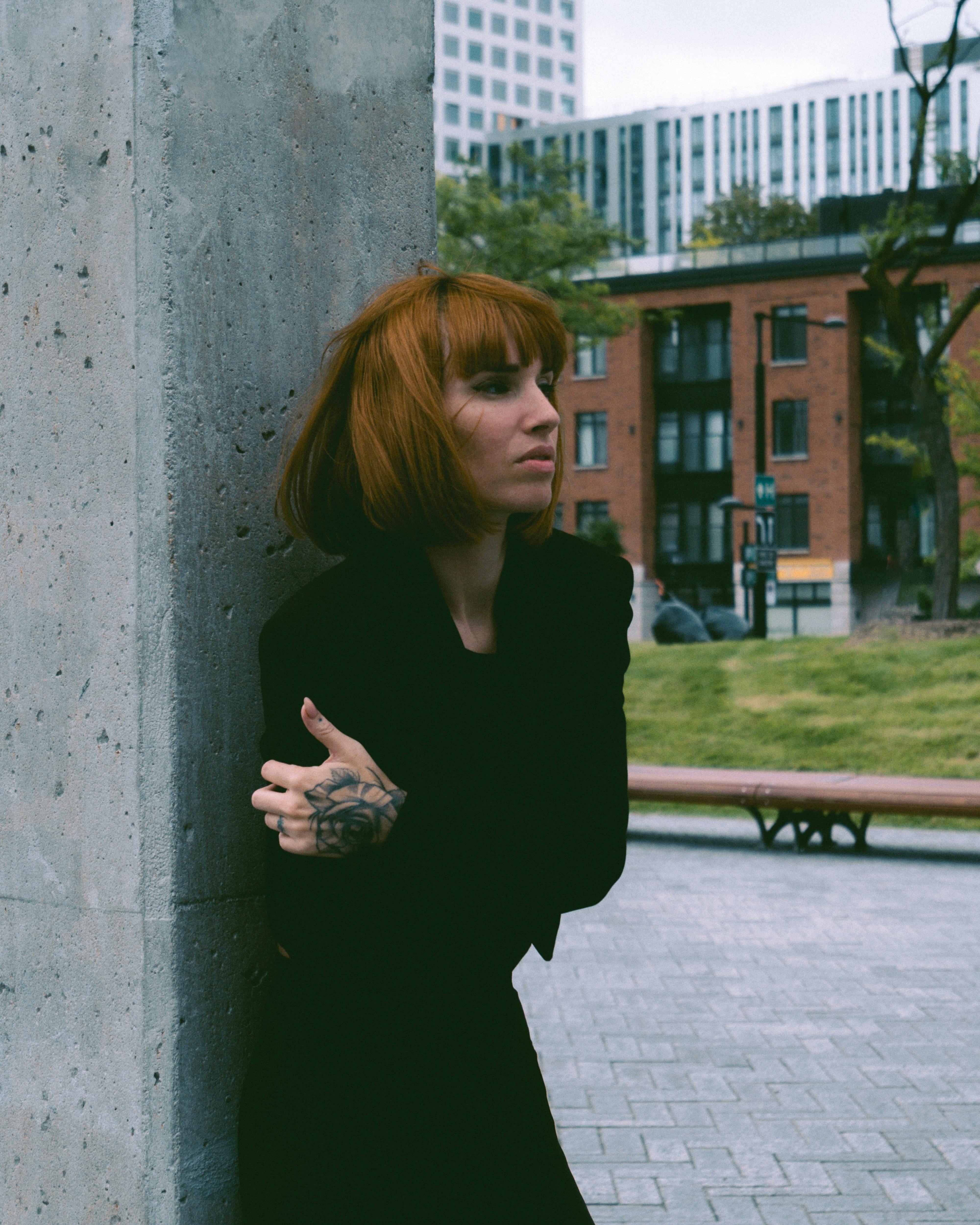 Creative urban portrait of a red-haired woman in a black outfit leaning against a concrete wall.