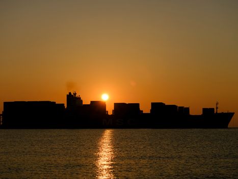 Silhouette of a cargo ship at sunset, casting reflections on the calm ocean water.