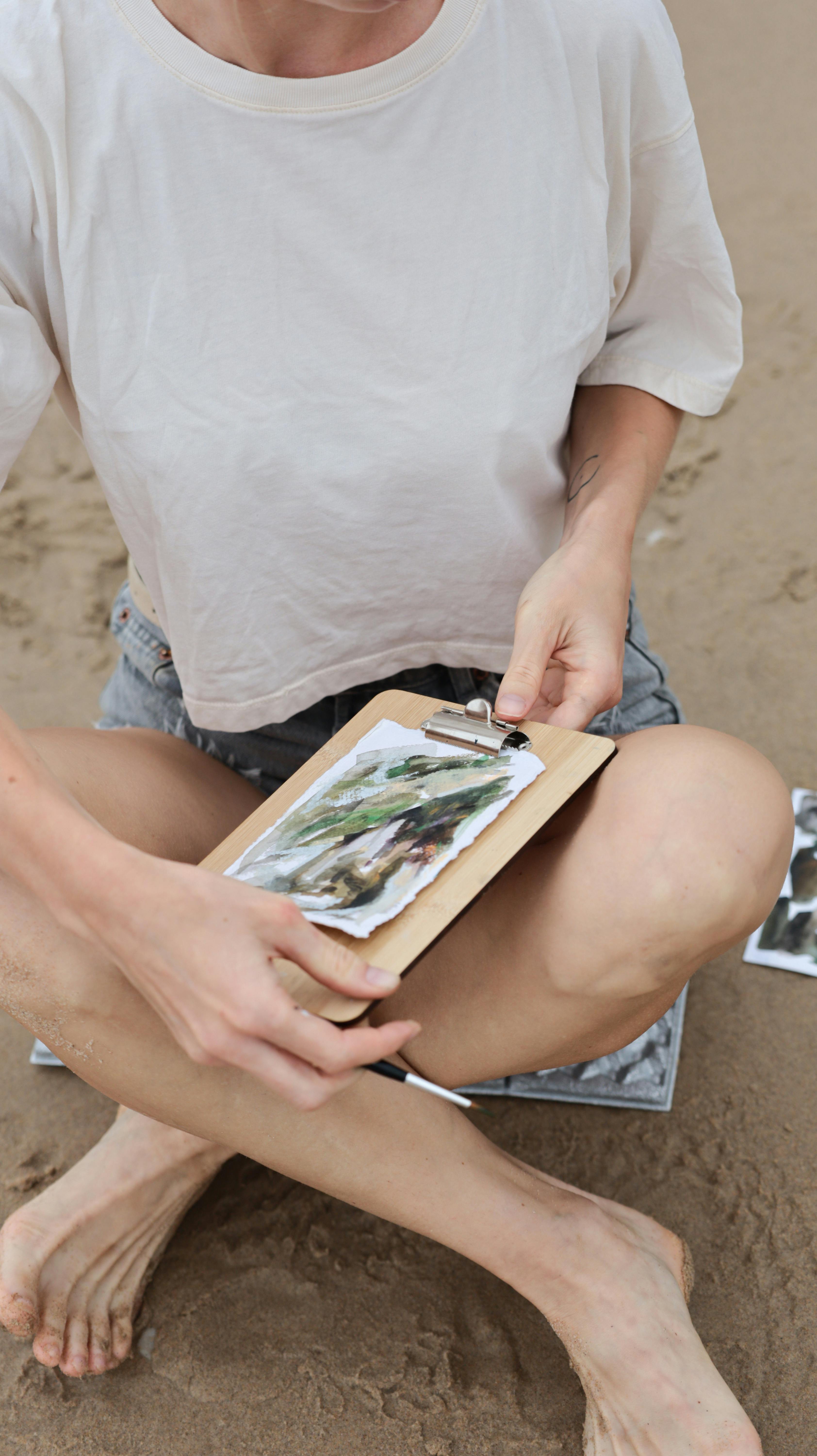 A woman sits on the sand drawing a beach scene, portraying creativity in summer outdoors.
