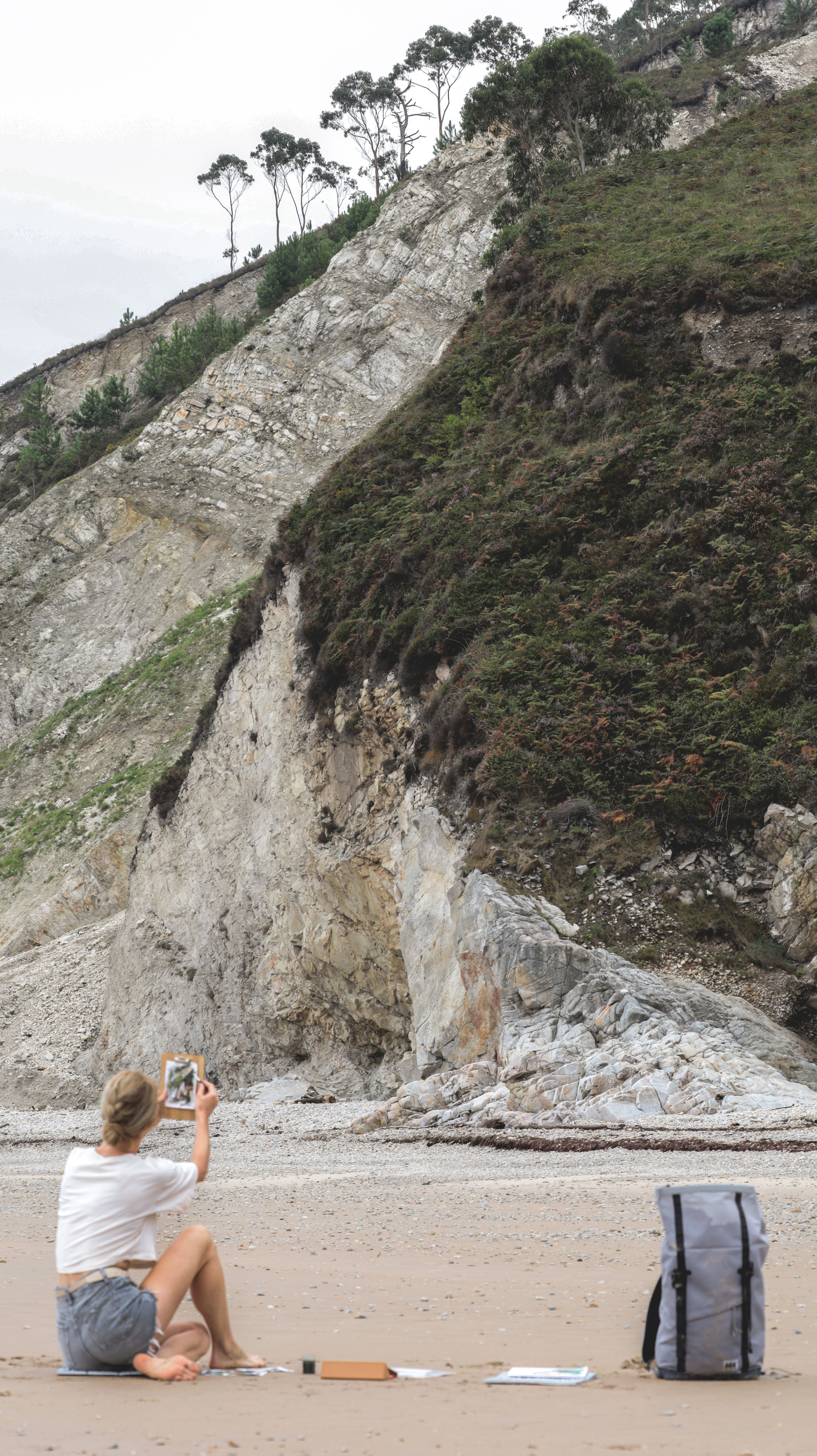 A woman captures the scenic view of a rocky beach with cliffs using her smartphone.
