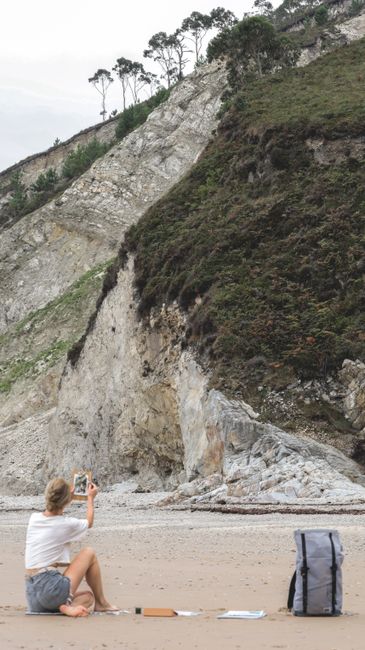 A woman captures the scenic view of a rocky beach with cliffs using her smartphone.