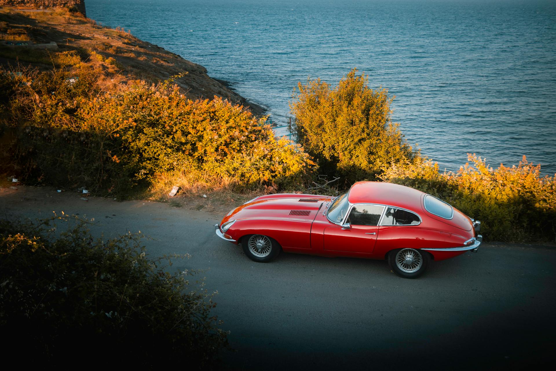 Classic red Jaguar E-Type Series 2 parked on a coastal road with scenic sea view.