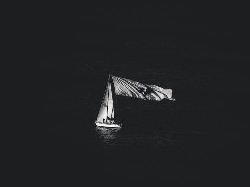 Free Black and white photograph of a sailboat with a large flag on the ocean at night. Stock Photo