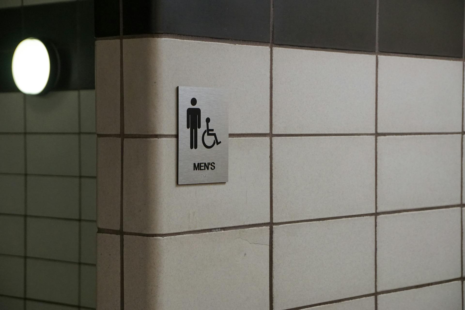 A men's restroom sign with accessible symbol on a tiled wall, featuring minimalist design.