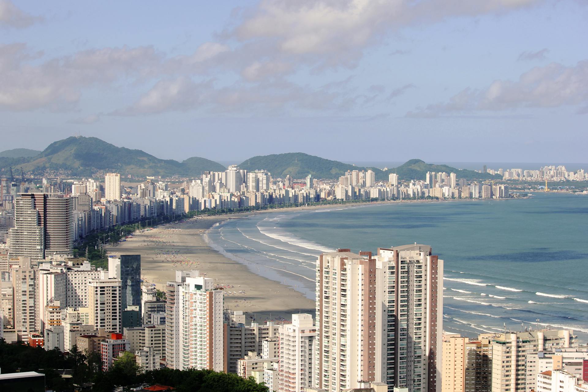 A panoramic view of Santos, Brazil showcasing skyscrapers along the coastline with ocean waves and nearby hills.