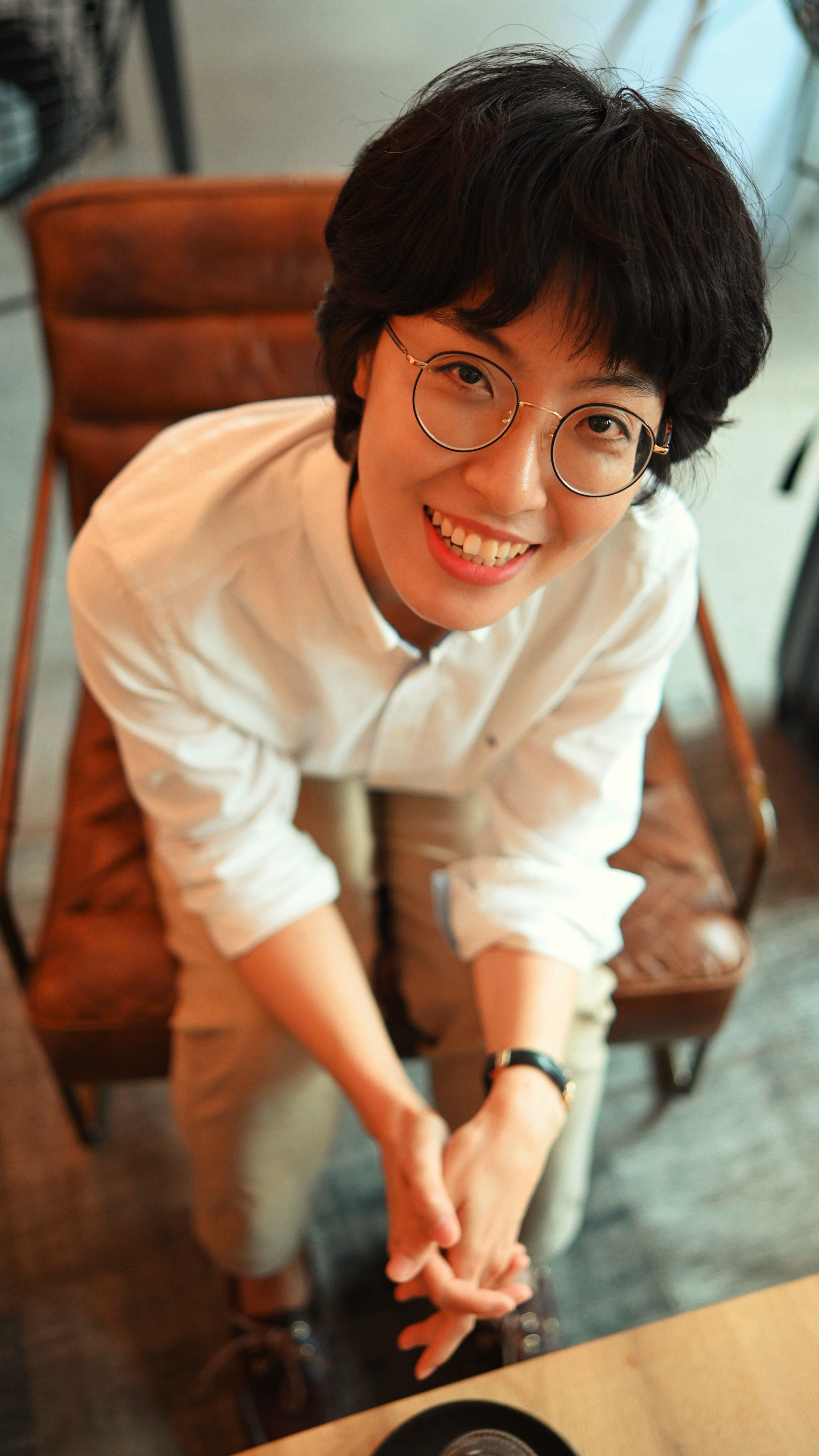 Happy woman wearing glasses and white shirt, sitting in a café, looking up with a smile.