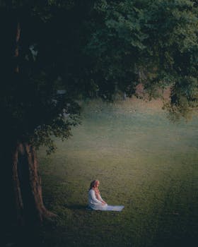 A woman in white dress sitting under a tree in a lush park, embodying tranquility and reflection.