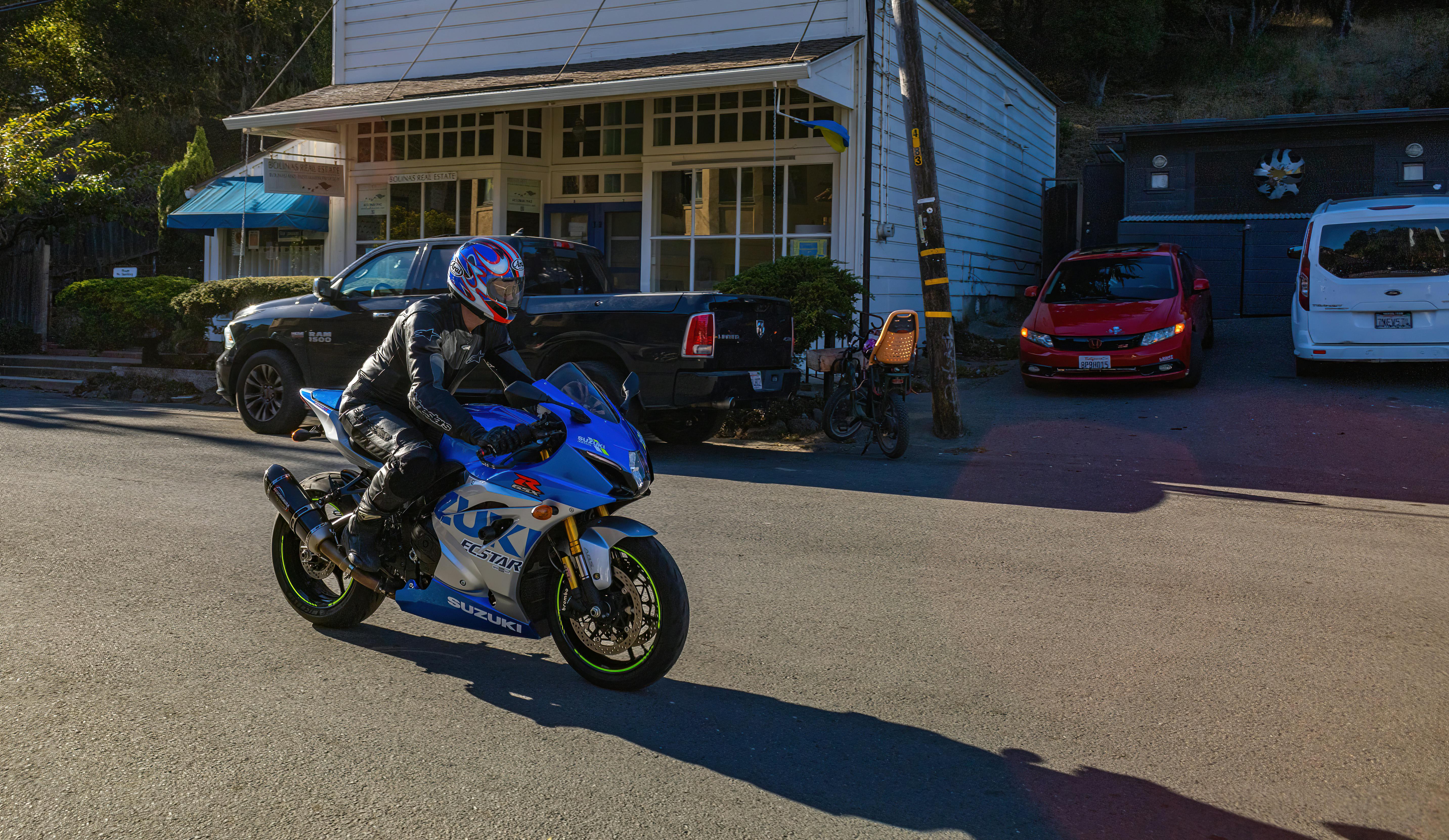 A motorcyclist in gear rides a blue sports bike on a sunny street.