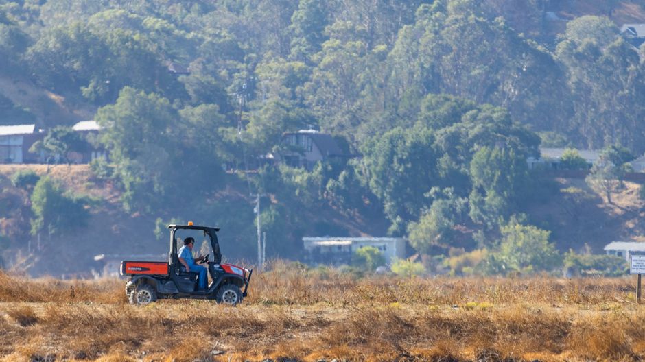 A man driving a utility vehicle through a rural farm with hills and trees in the background.
