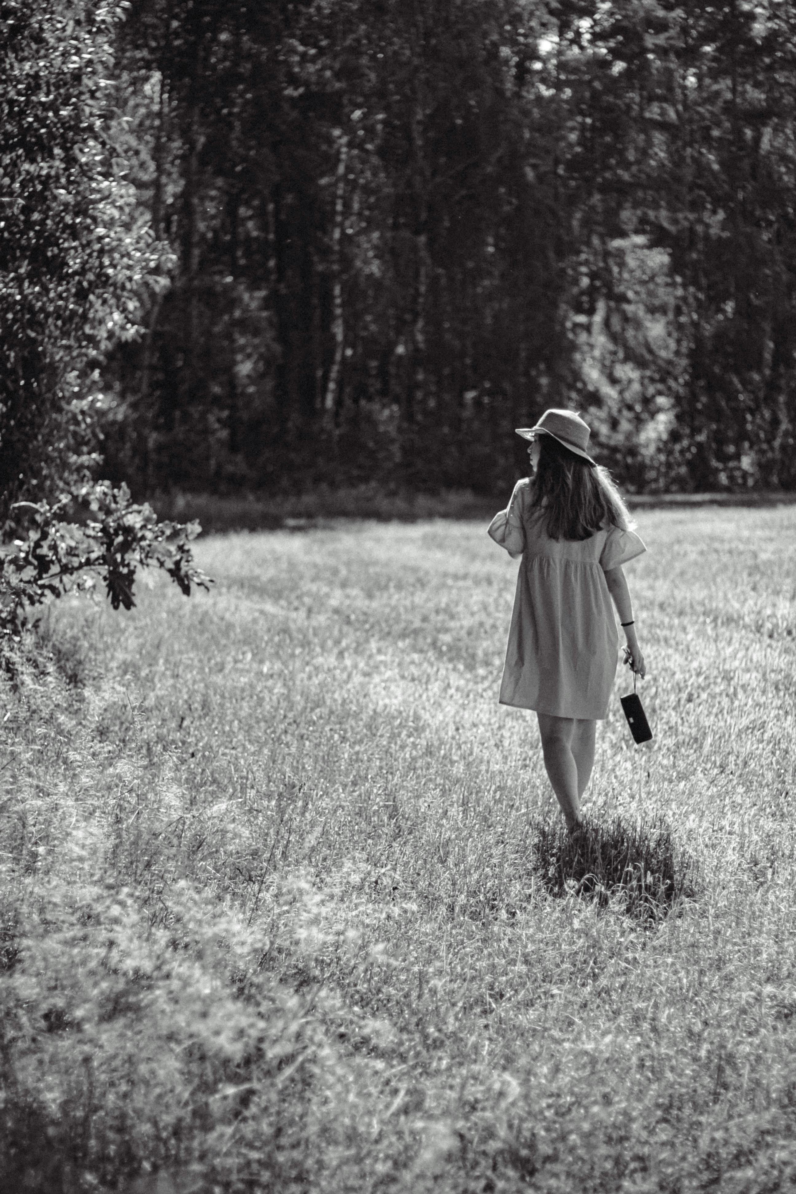 A woman in a summer dress and hat walks through a sunlit grassy field with trees.