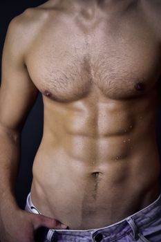 Close-up of a muscular male torso with defined abs and well-toned body, shirtless against a dark background.