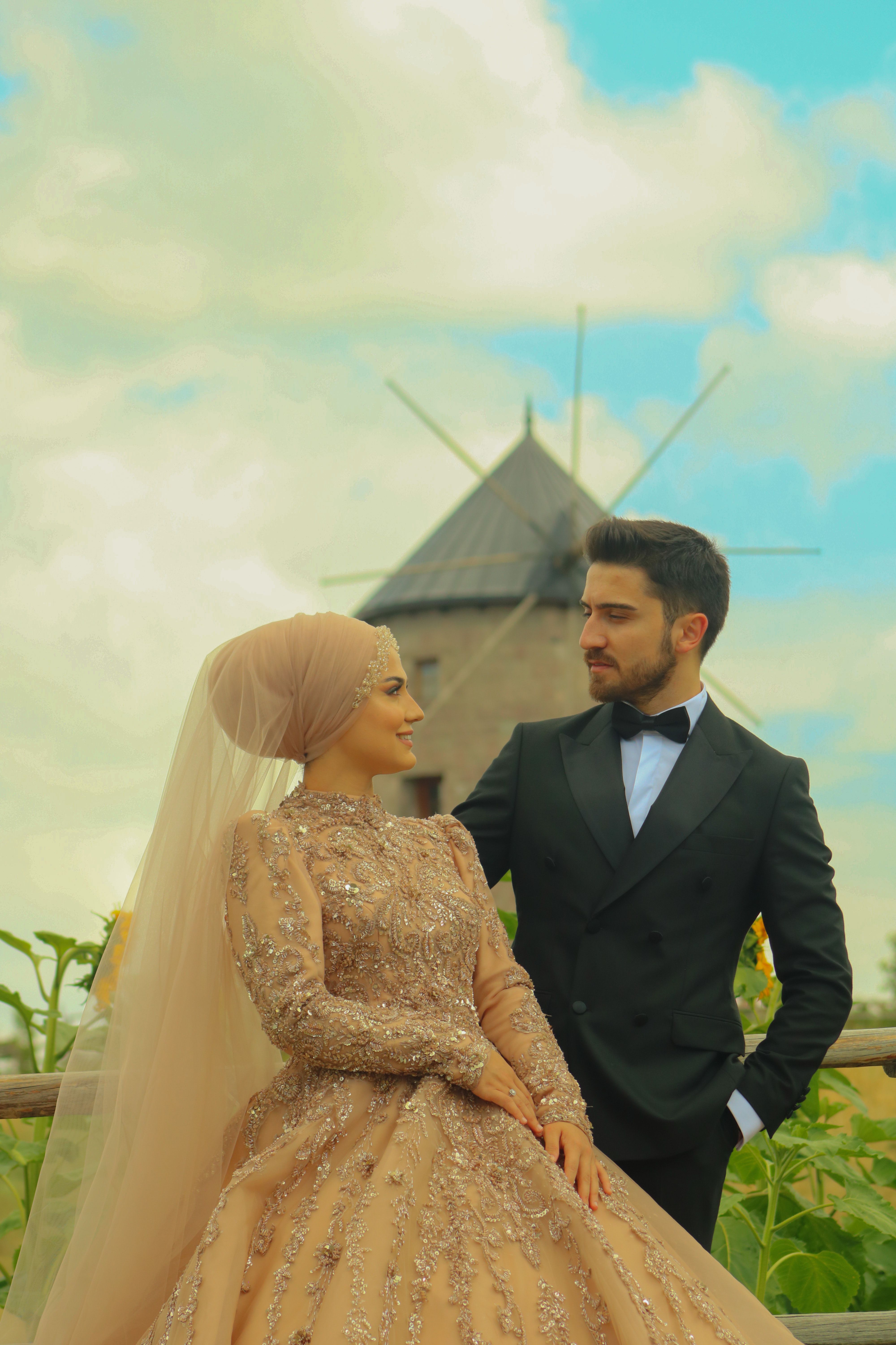 Charming wedding couple poses in elegant attire beside a rustic windmill under a blue sky.