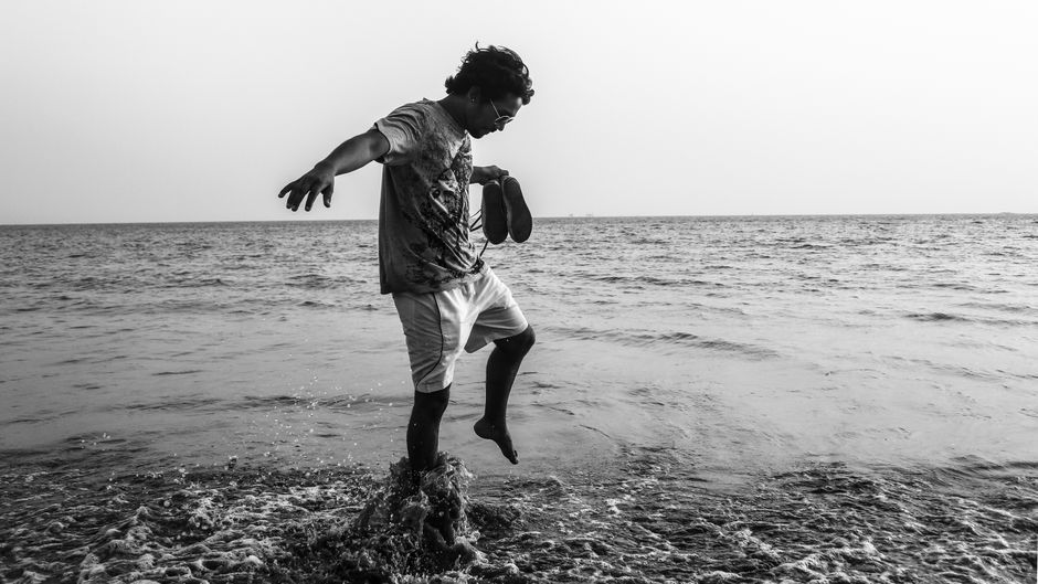 A man holding shoes jumps joyfully on a beach, captured in monochrome.