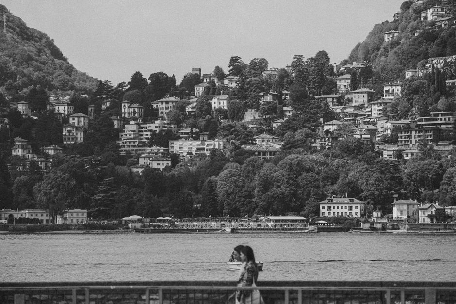 Black and white image of a hillside village with a lake and a woman walking, conveying a peaceful atmosphere.