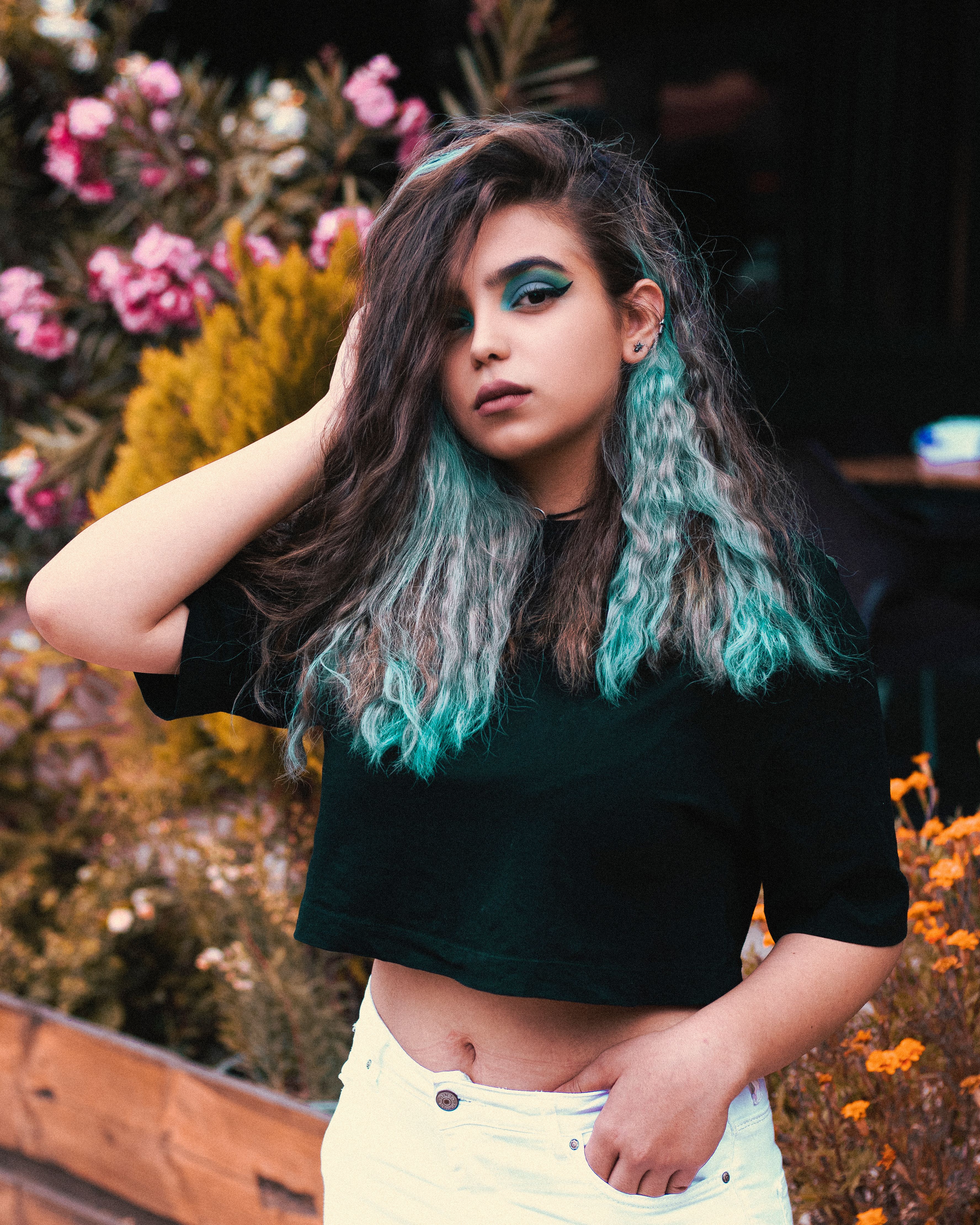 Fashion portrait of a stylish young woman with blue hair standing outdoors amidst colorful flowers.