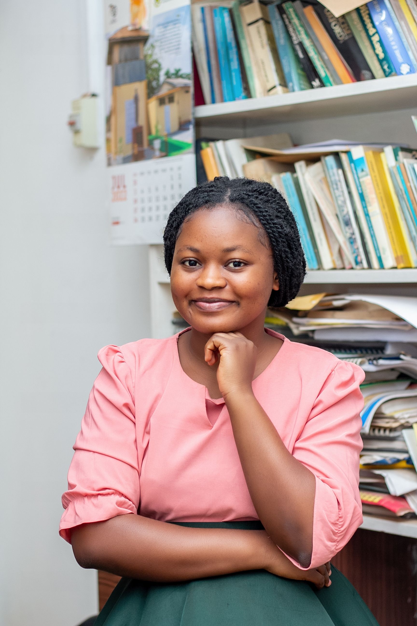Smiling woman with braids in pink, posing thoughtfully in front of a bookshelf.