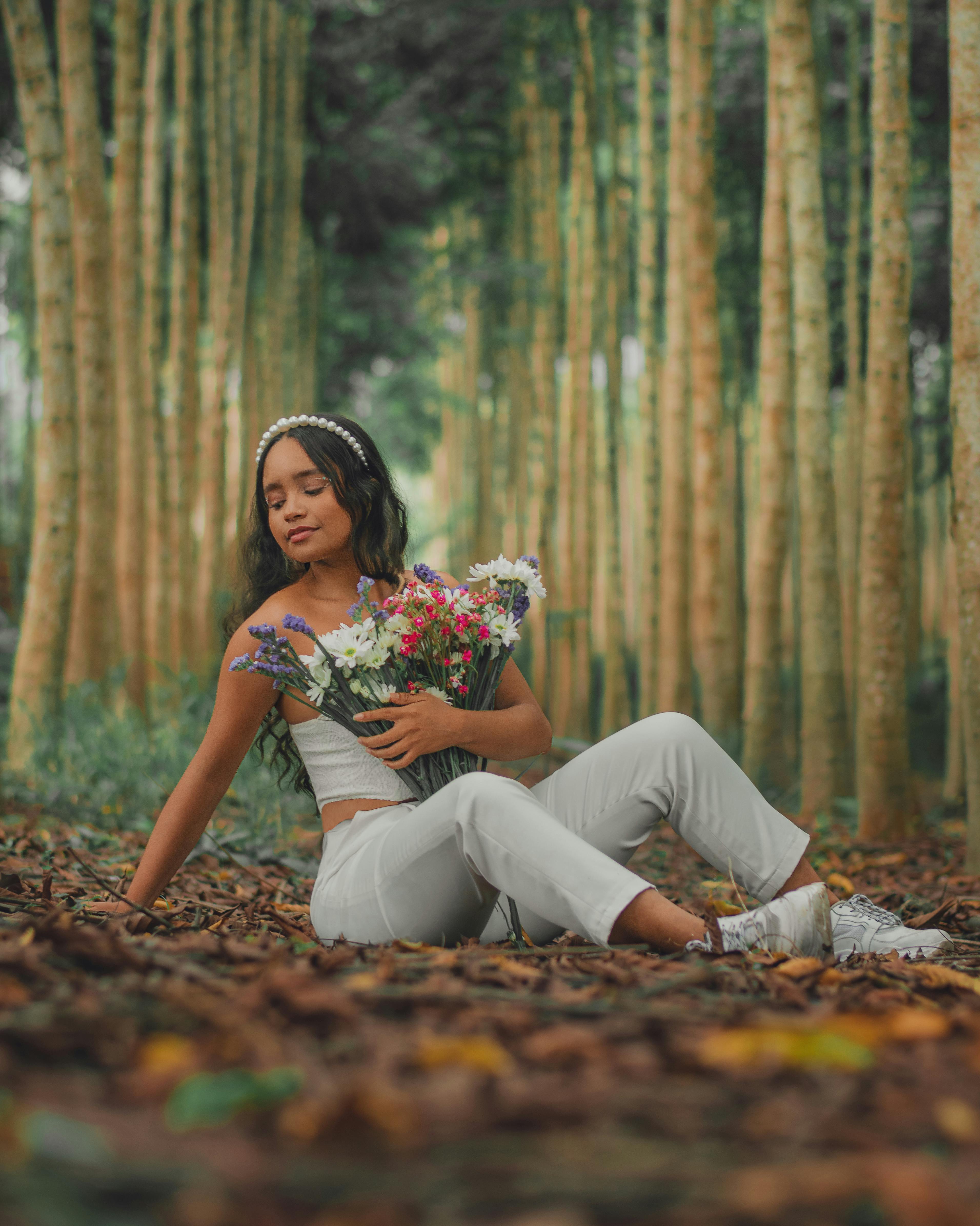 A woman in a forest sits holding flowers, surrounded by autumn leaves, creating a serene and relaxed atmosphere.