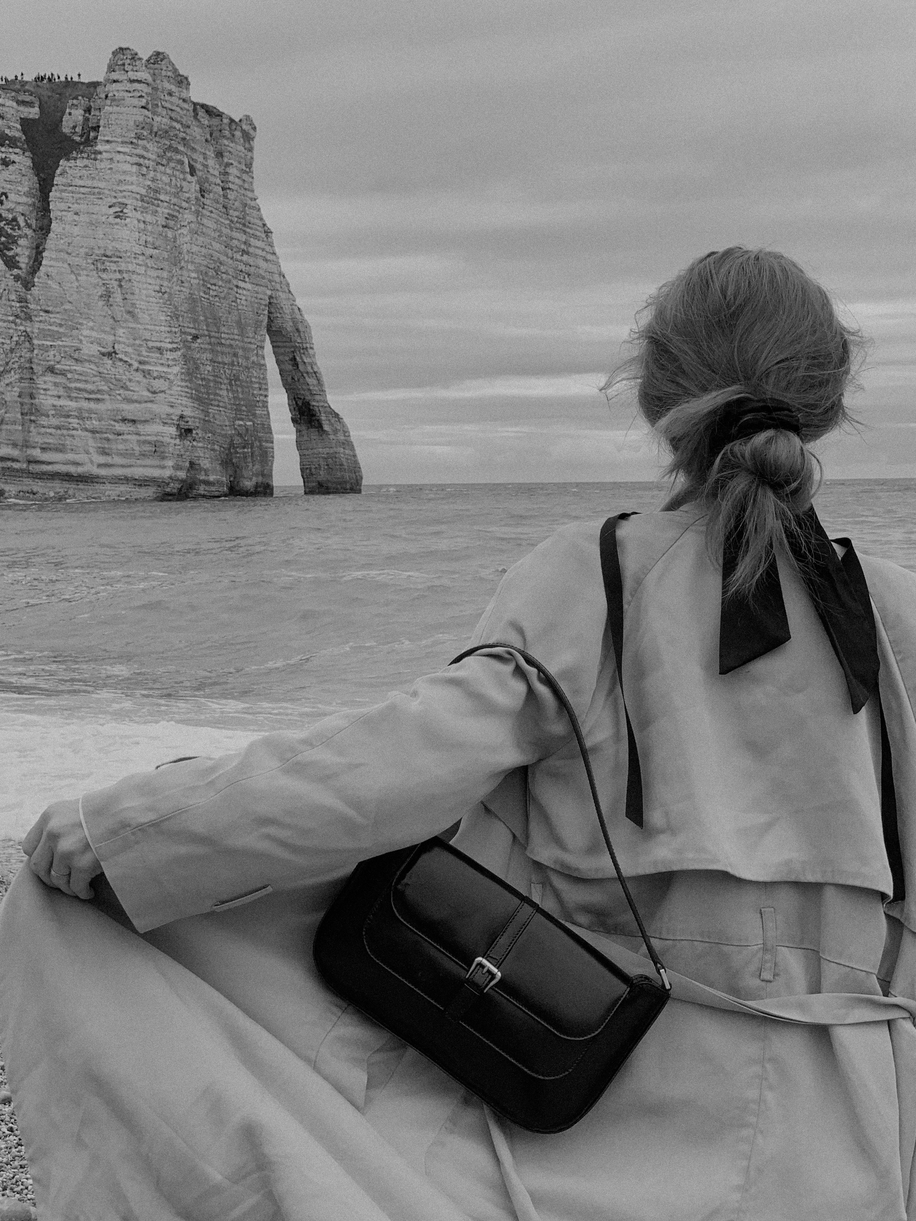 A woman stands on a beach with a handbag, facing a sea cliff in a black and white photograph.