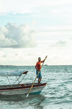 Man in a red shirt paddling a small boat under a cloudy sky on the sea.