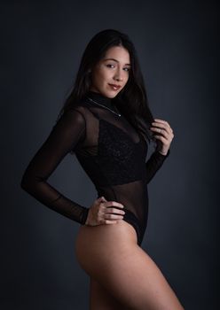 Side profile studio portrait of an elegant woman in a sheer black bodysuit with hand on waist.