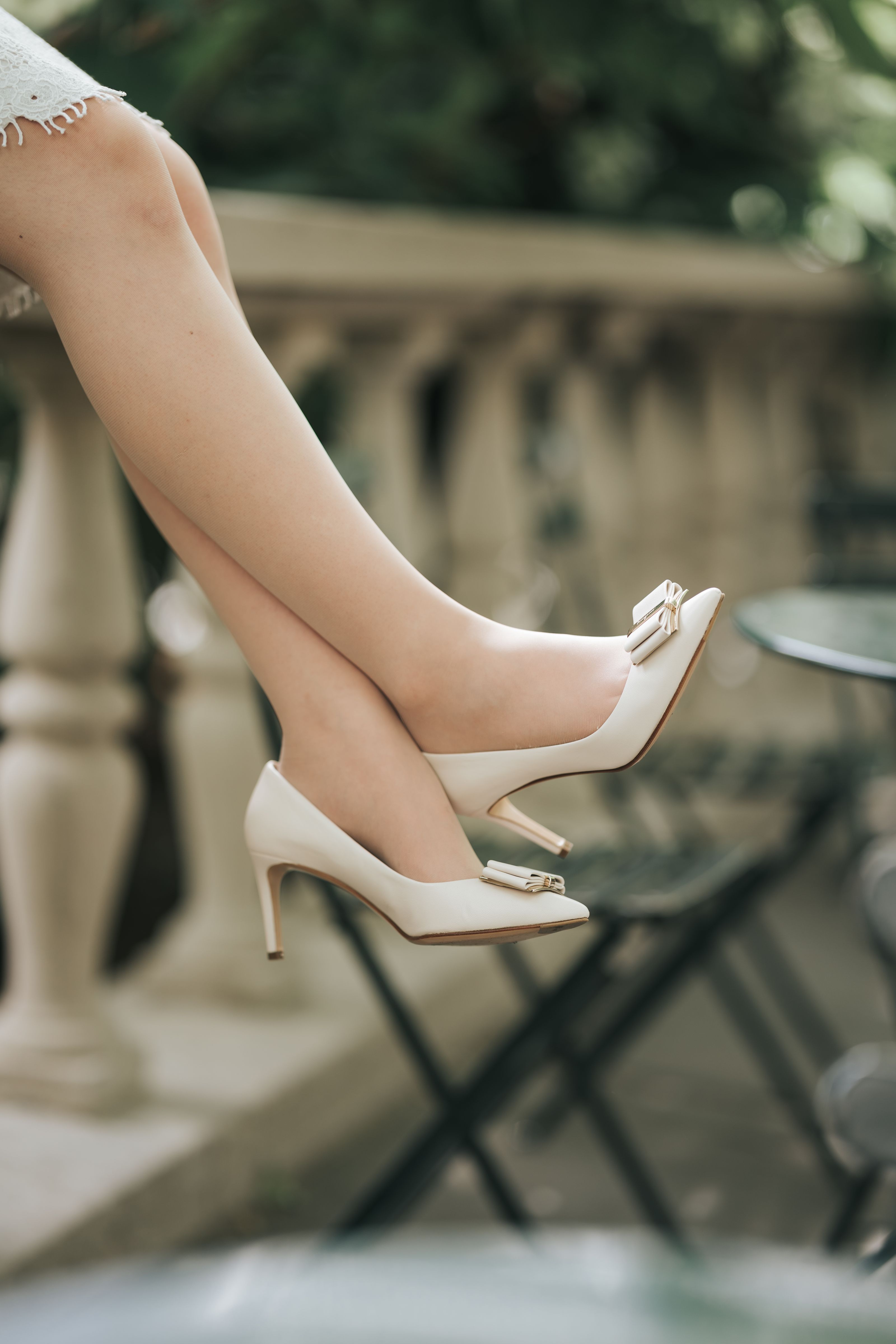 Close de sapatos brancos de salto alto com laços e pernas cruzadas ao ar livre, transmitindo sofisticação.