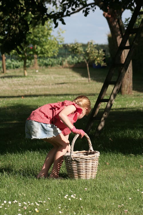 Girl in Red Shirt and Blue Denim Shorts Standing on Green Grass Holding Brown Woven Basket
