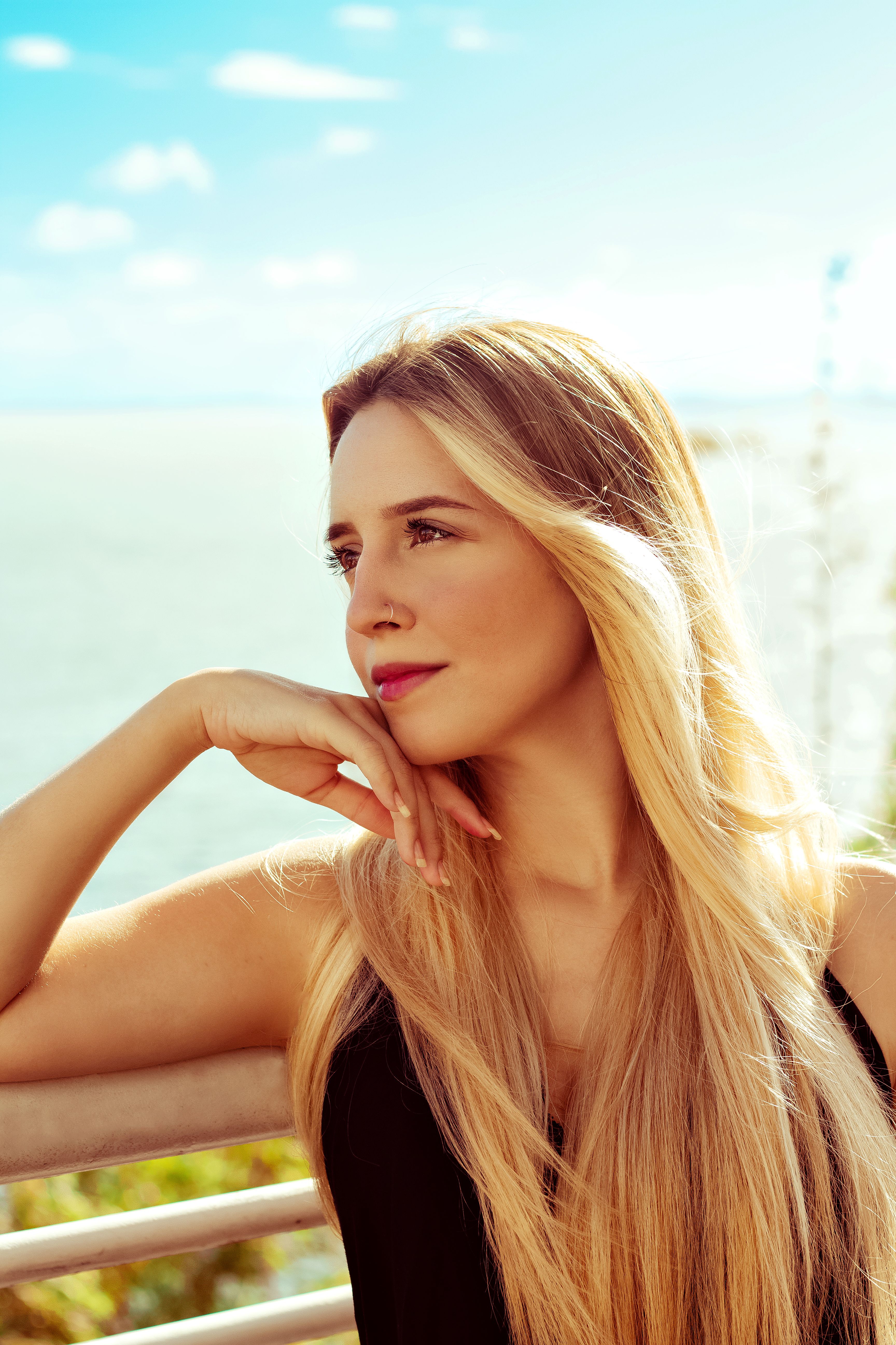 Fotomural Retrato de una mujer rubia serena junto al mar, con la mano en la barbilla y una expresión pensativa.