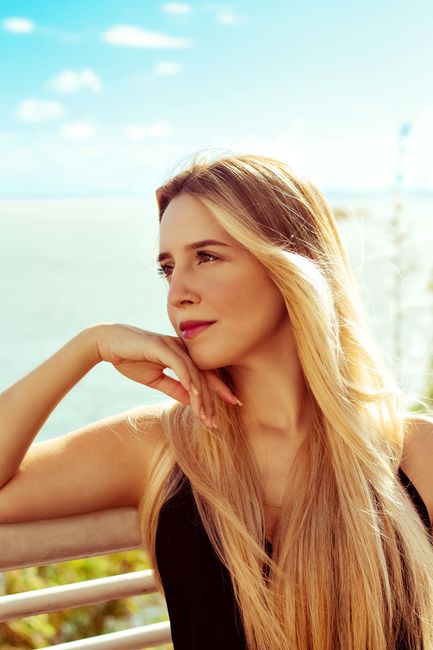 Fotomural Retrato de una mujer rubia serena junto al mar, con la mano en la barbilla y una expresión pensativa.