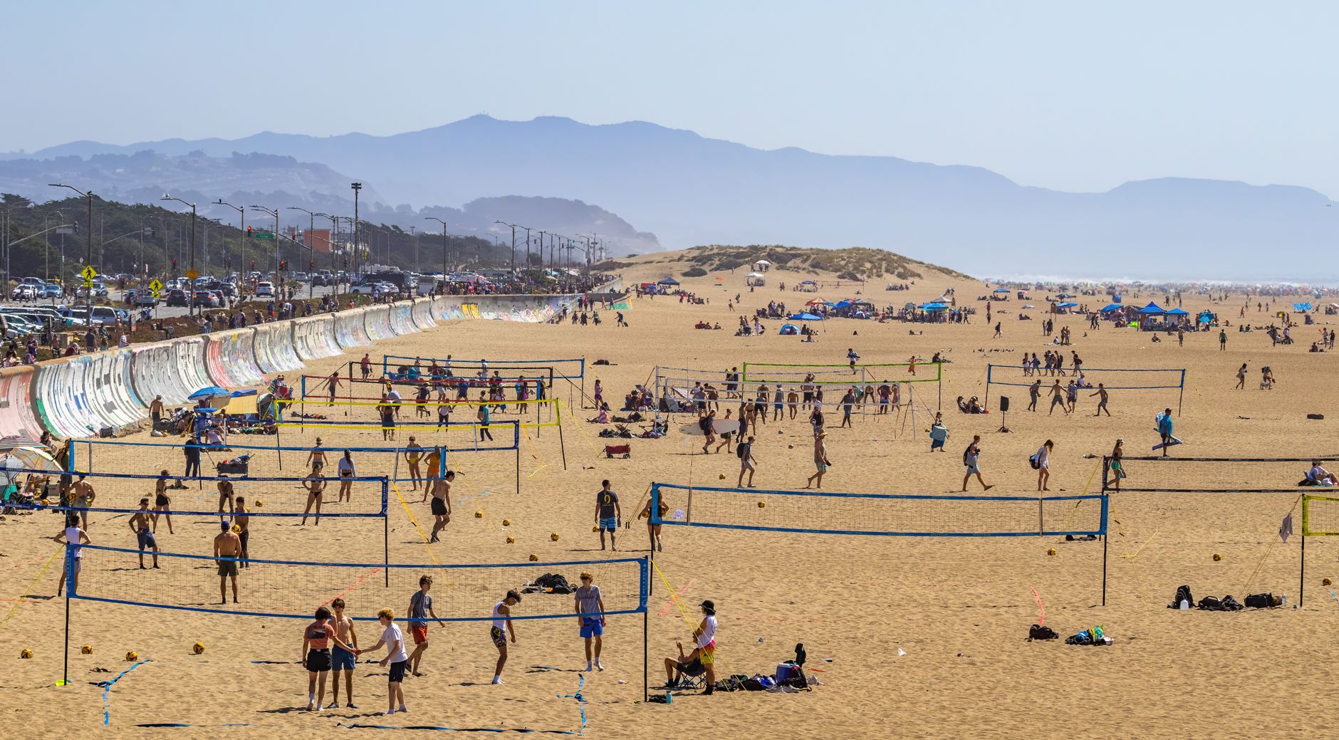 Aerial view of multiple beach volleyball games on a sunny day at a busy coastal beach.