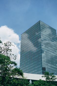 A modern glass skyscraper reflects a blue sky, surrounded by greenery.