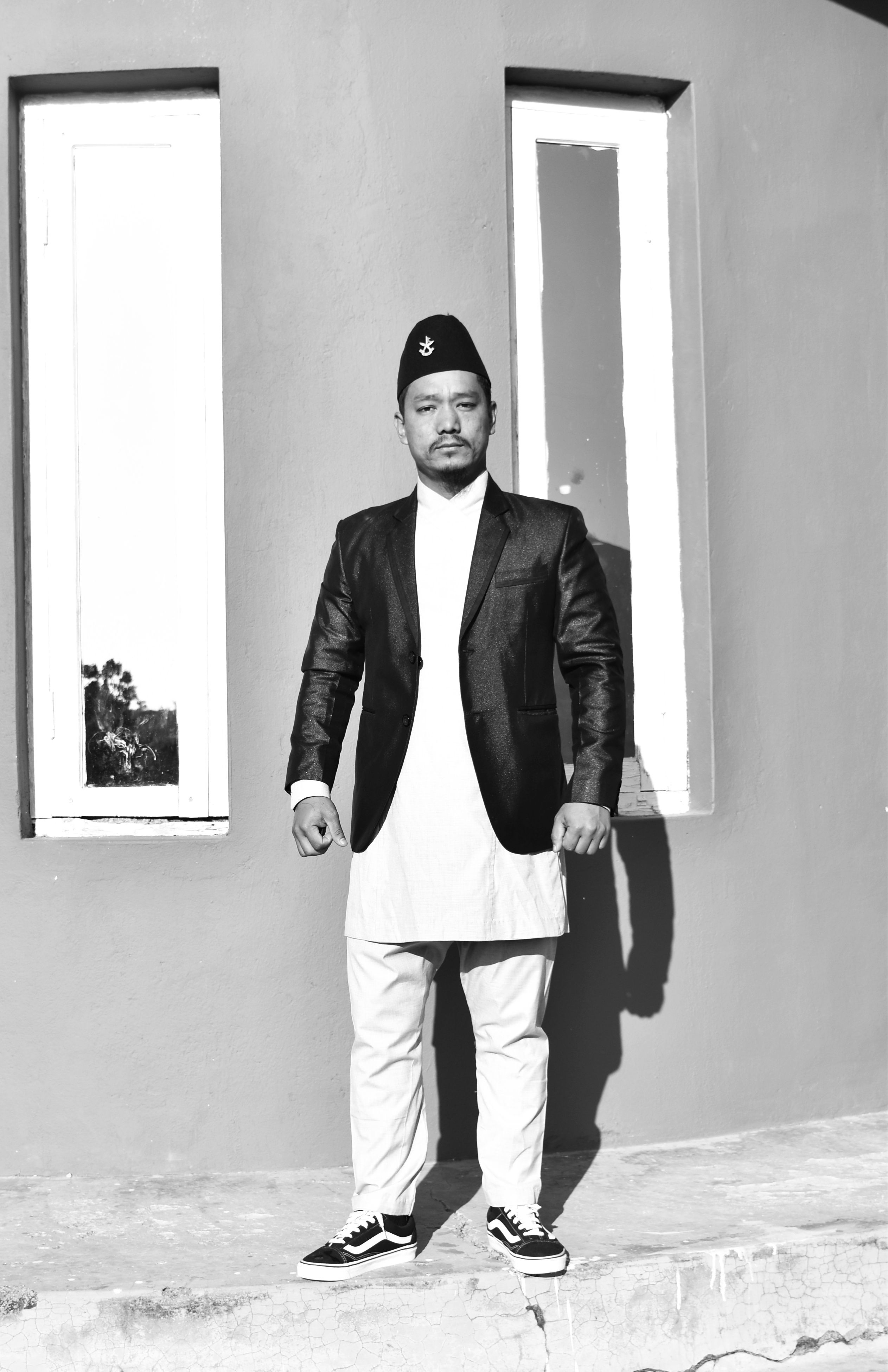 Man in a traditional outfit and cap stands by a concrete wall in a black and white portrait.