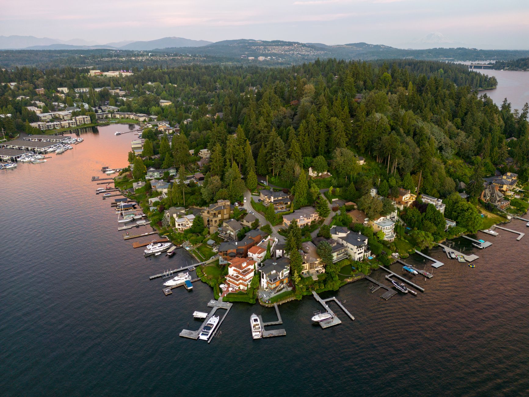 Scenic aerial view of waterfront homes, marina, lush greenery, and distant mountains during sunset.