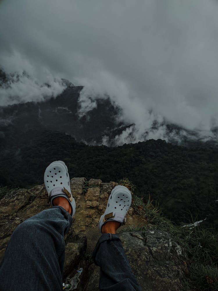 Person In Blue Denim Jeans And Clogs Sitting On A Cliff