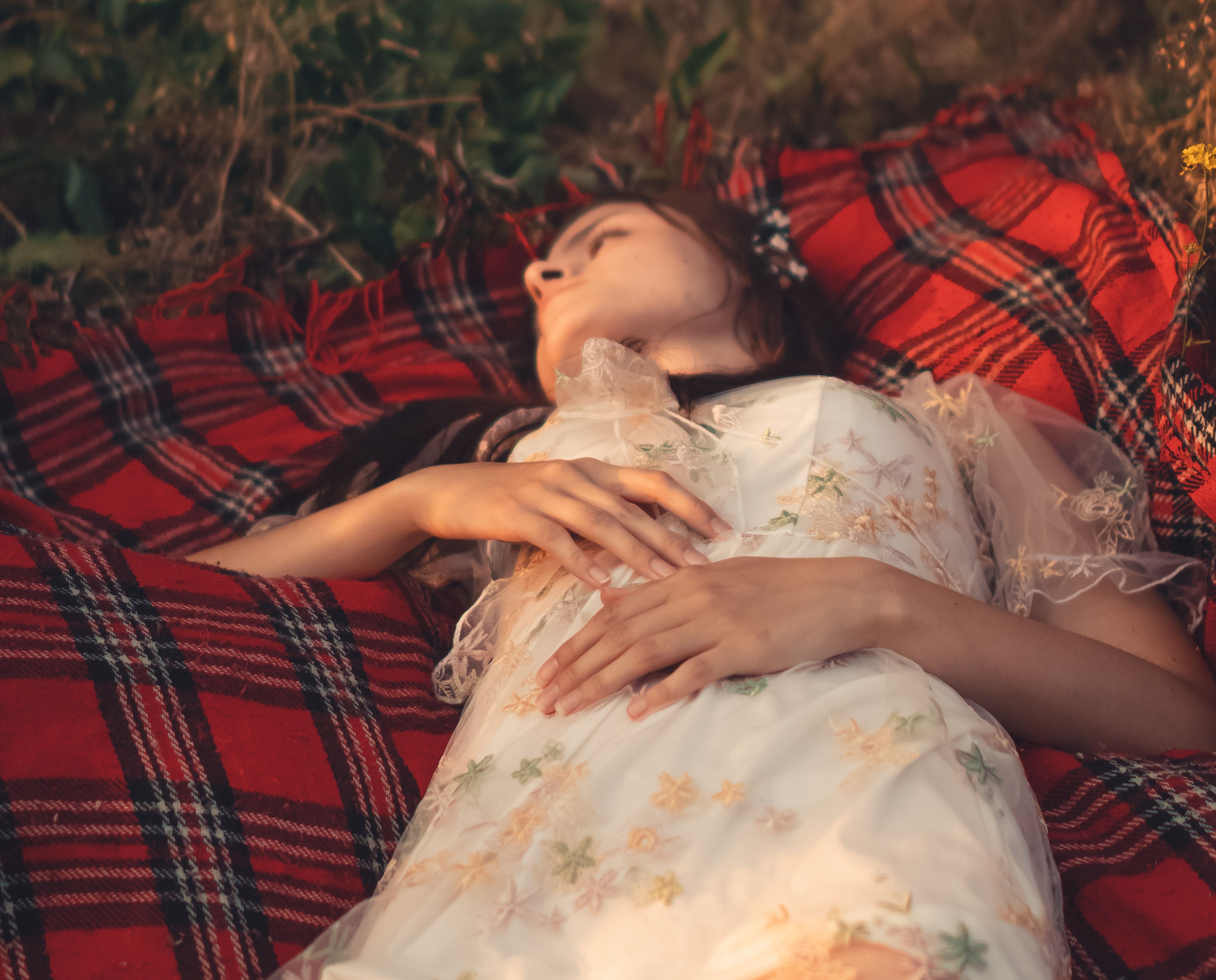A young woman in a floral dress relaxing on a plaid blanket outdoors during summer.