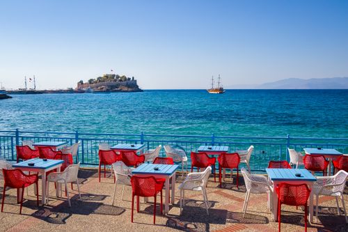 Free Outdoor seaside cafe with vibrant red chairs, blue tables, and a clear view of a distant island and sailing boat. Stock Photo