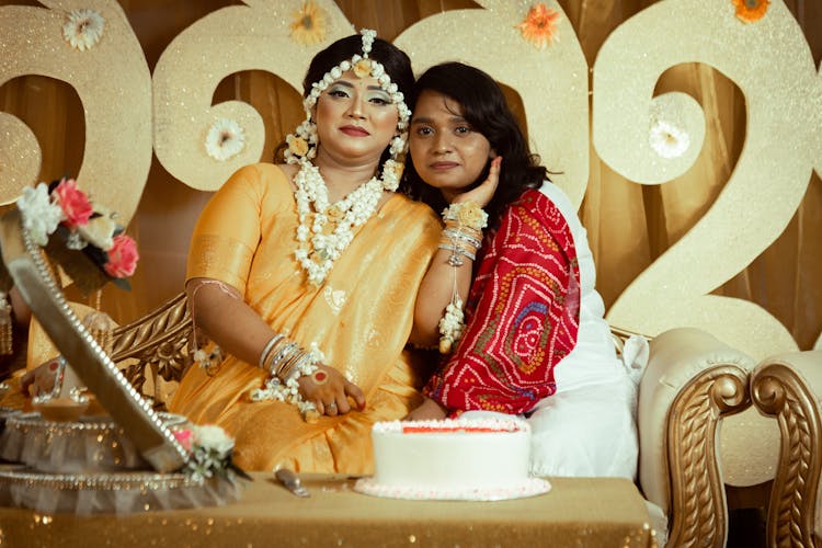 Beautiful Bride In Her Wedding Dress Posing Beside Her Sister Sitting On A Sofa