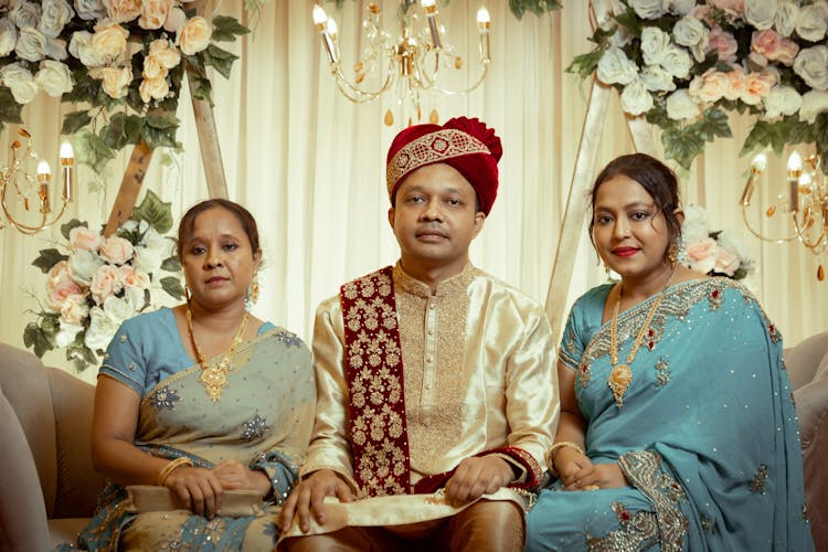 Groom In His Traditional Wedding Sherwani Suit Sitting On A Sofa Between His Sisters In Elegant Saree Dresses